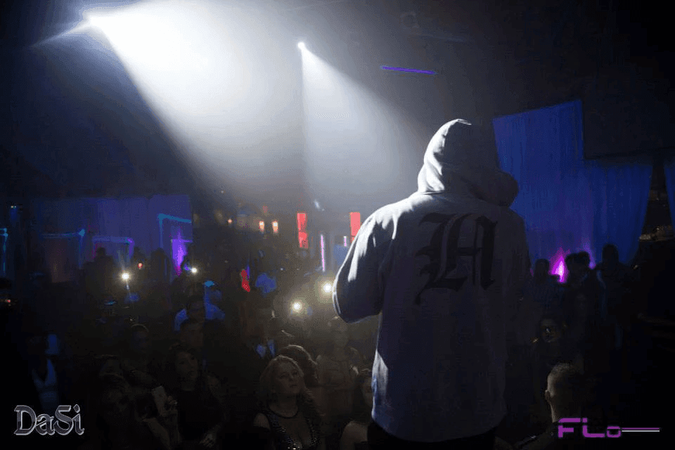 Performer in hoodie faces a packed club crowd under hazy white beams