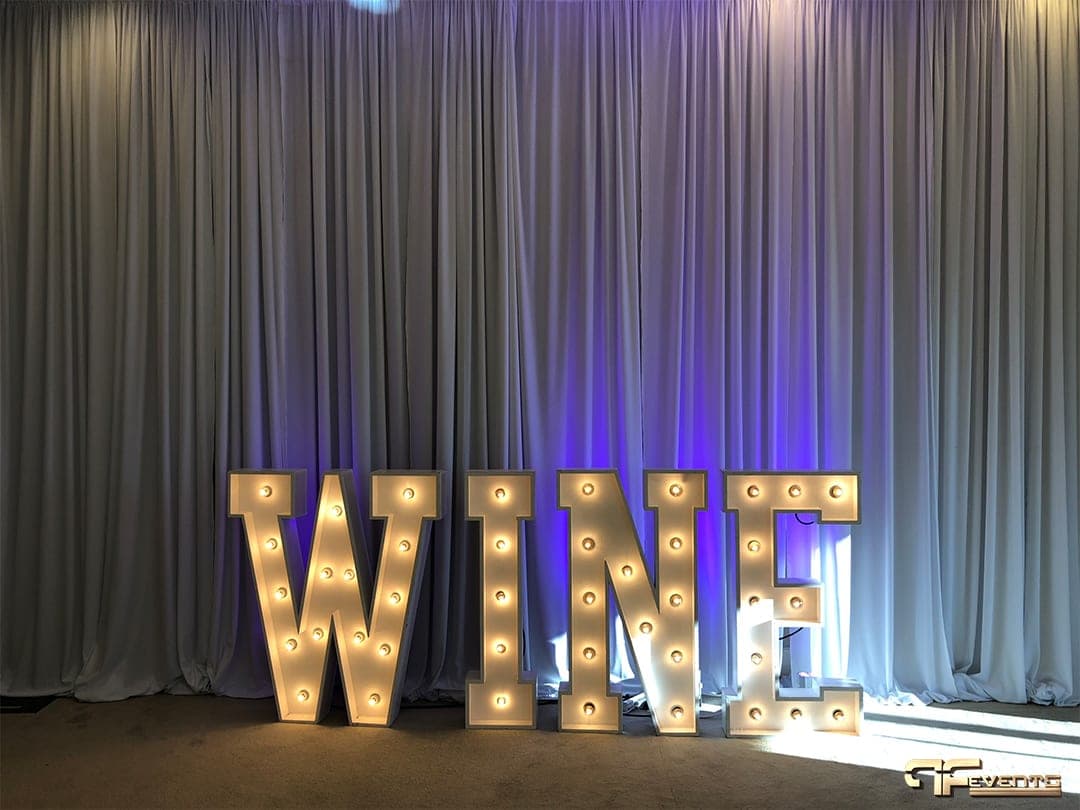 Light-up marquee letters spelling WINE before white drape with purple uplight