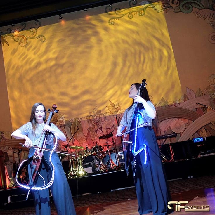 Electric cello duo beneath a gold water-ripple light effect on the proscenium wall