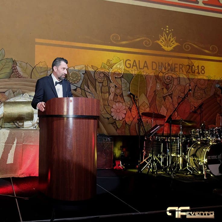 Speaker at a wood podium below a projected Gala Dinner 2018 banner and band stage