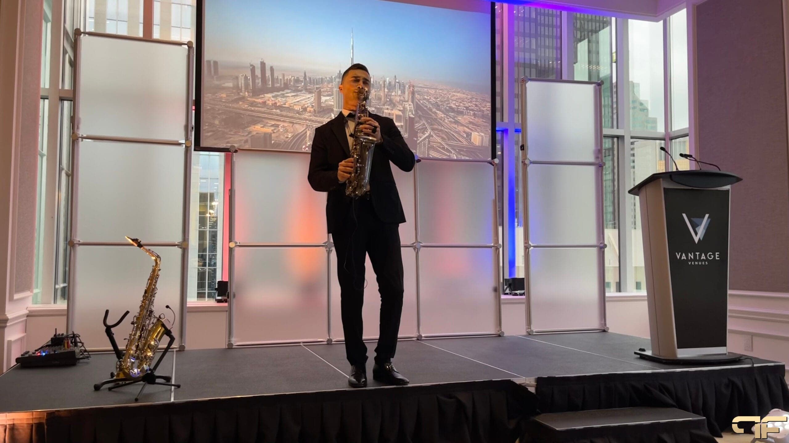 Saxophonist on the black stage deck, Dubai skyline on screen, Vantage lectern at right